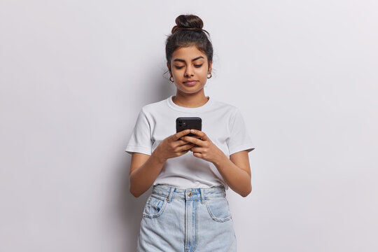 Pretty young Indian woman engaged in modern communication poses with her smartphone in hand chats online connecting with others wears casual t shirt and jeans isolated over white background.