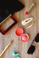 Handbag, sunglasses, beauty products, pearl hair clips, rings, pen, earbuds, book and phone on wooden background. Top view.