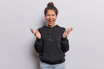 Emotional dark haired Latin woman keeps palms raised up exclaims loudly has eyes closed wears black hoodie and jeans isolated over white background. People negative emotions and reactions concept
