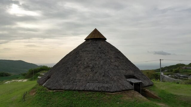Rise and pullback over historic thatched-roof hut on grassy hill by village
