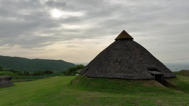 Rotating around historic Jomon thatched-roof hut on grassy hill 
