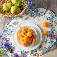 Summer mood: fresh apricots, pears and cornflower flowers on a wooden background surrounded by vintage crockery. Top view - 612047622