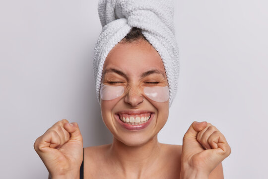 Headshot of beautiful young woman dedicates moment to self care gently places collagen beauty patches under eyes to reduce wrinkles smiles toothily clenches fists poses with bath towel wrapped on head