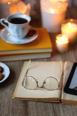 Cup of tea or coffee, plate of cookies, books, e-reader, pencil and lit candles on the table. Selective focus.