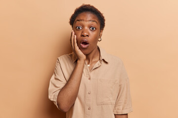 Dark skinned African woman gazes at camera with wide eyes and an open mouth her hand cheek reacts with astonishment on something horrible wears shirt isolated over brown background. Omg concept
