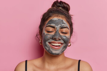 Headshot of cheerful beautiful woman adorns her face with charcoal black mud mask personifies essence of beauty and healthcare keeps eyes closed stands bare shouldered isolated over pink background