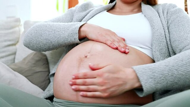 Woman sitting on a sofa at home, gently applying skin care cream to her belly in the last months of pregnancy to prevent pregnancy stretch marks. Pregnancy third trimester - week 34.