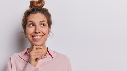 Studio shot of beautiful woman smiles toothily has gentle expression smiles tenderly keeps hand under chin concentrated wears shirt isolated on white background copy space for your advertising content