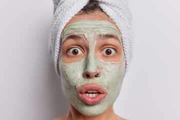 Young woman undergoes spa procedures applying facial beauty mask isolated over white background delves into world of skin care seeking to rejuvenate and enhance complexion has bath towel on head