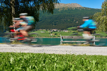 Obraz na płótnie Canvas 3 cyclists in motion. Adults doing sports with bicycle on sunny summer afternoon, at the lake in the mountains. Italy, Vinschgau, Lake Resia (Reschensee).