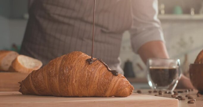 Dark chocolate is poured onto a french croissant with chocolate filling on table with cup of coffee, french baguette and coffee beans