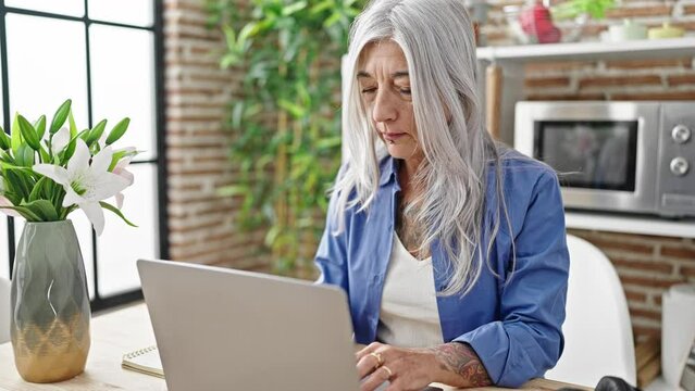 Middle age grey-haired woman closing laptop stretching arms at dinning room