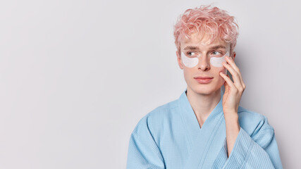Horizontal shot of pink haired man highlightes significance of beauty and wellness applies beauty patches under eyes to reduce wrinkles wears blue robe isolated over white background copy space