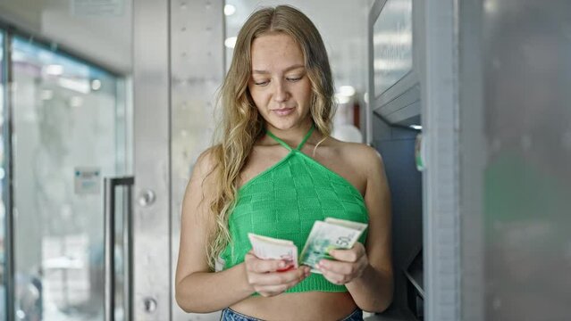 Young blonde woman holding israel shekels banknotes of cash machine at street