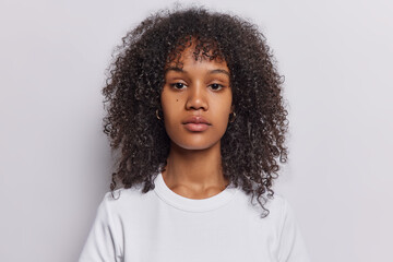 Portrait of serious curly haired woman looks directly at camera has attenitve gaze calm expression dressed in casual t shirt isolated over white background. Pretty millennial girl poses in studio