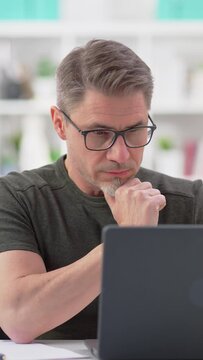 50s businessman with gray hair working from home, thinking. Man in casual sitting at desk using laptop computer, business manager online in home office.
