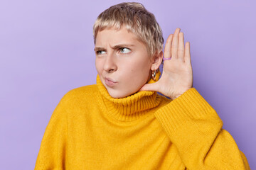 Displeased short haired woman frowns face keeps hand near ear tries to overhear information listens...
