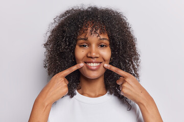 Headshot of pretty curly haired woman points at toothy smile demonstrates perfect teeth dressed in casual t shirt isolated over white background. People positive emotions and feelings concept