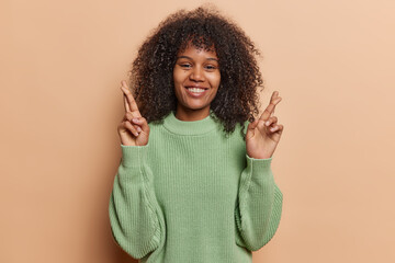 Pretty curly African woman stands with crossed fingers warm smile emanating strong belief in positive outcomes her faith in good fortune reflects spirit of hope resilience wears casual knitted jumper - obrazy, fototapety, plakaty