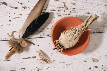 Smudge stick with White Sage (Salvia apiana) in a clay bowl and incense feather on old white wood. Overhead view.
