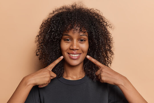 Photo of lovely curly haired African woman points fingers at toothy smile shows white perfect teeth dressed in casual black t shirt isolated over brown background. Look at my brilliant smile