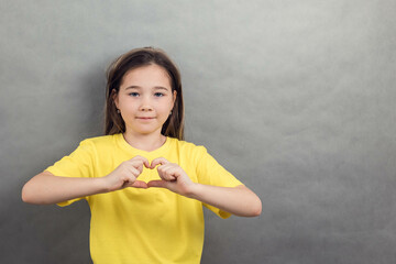 Close-up portrait of an Asian girl in a yellow T-shirt on a gray background, making a heart with her hands, copy space