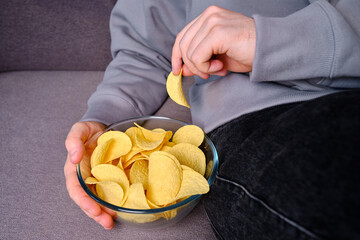 A man eats crispy potato chips from a transparent bowl on the couch. Quick snack. Calories and...