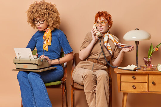 Two busy women in old fashioned attire diligently work on typewriter and engage in phone call using stationary telephone surrounded by paperwork. Nostalgia timeless elegance. Vintage inspired scene