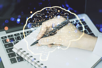 Creative artificial Intelligence concept with human brain hologram and hand writing in notebook on background with laptop. Multiexposure