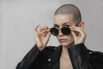 Portrait of a beautiful young woman with a shaved bald head in dark sunglasses and a black leather jacket on a light background
