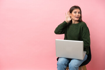 Young caucasian woman sitting on a chair with her laptop isolated on pink background listening to something by putting hand on the ear
