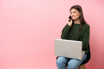 Young caucasian woman sitting on a chair with her laptop isolated on pink background keeping a conversation with the mobile phone with someone