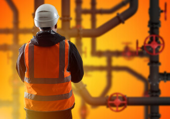 Factory worker. Man has back to camera. Industrial pipes behind guy. Factory worker in orange vest....