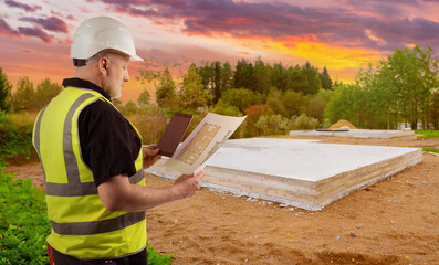 Man builder. Architect near foundation of house. Builder with architectural drawings. Man in work uniform holds clipboard. Builder in reflective vest. Guy foreman near forest with sunset