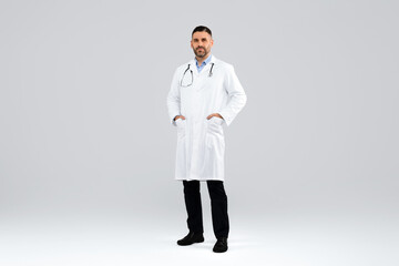 Confident middle aged doctor in workwear posing with hands in pockets on light background, full length