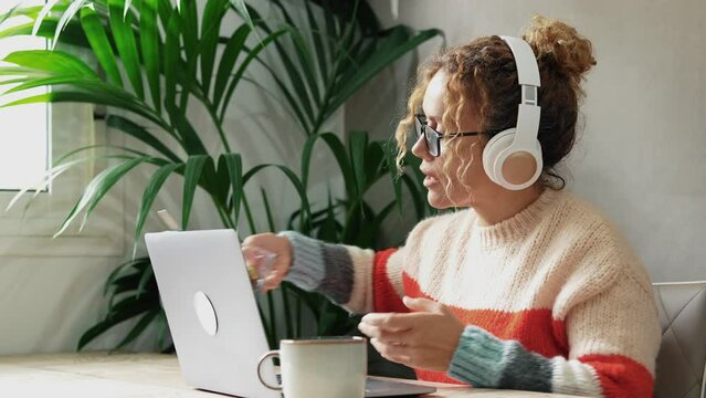 Happy young adult woman at work at home on laptop online job wearing headphones and eating snacks. Concept of lunch break during job. Freelance free lifestyle indoor. Female having fun telecommuting