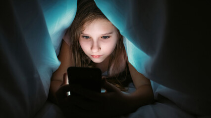 Child addiction. Online night. Focused serious obsessed girl lying hiding with smartphone under blanket watching content at bedtime at home.