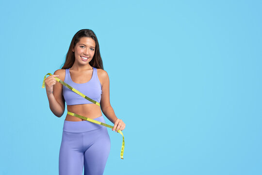 Smiling millennial caucasian slim woman in sportswear checking waist with measuring tape