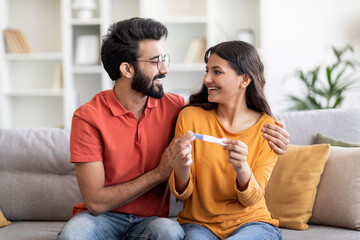 Happy indian couple holding positive pregnancy test and embracing at home - obrazy, fototapety, plakaty