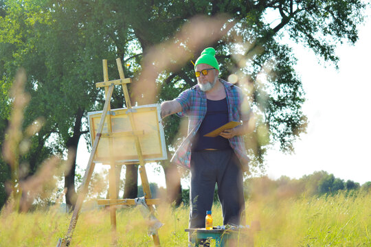Working Artisan painter creates picture with oil paints, applies strokes by paintbrush on canvas using wooden easel while creating artwork outdoors in dawn. Blurred meadow herbs grass on foreground