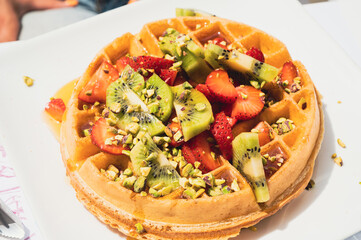 Large waffle with fresh strawberries, kiwi slices, pistachio pieces and honey on white plate