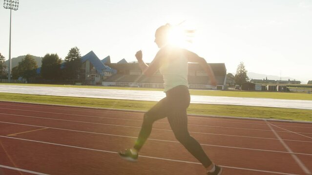 Young overweight woman running and jumping over hurdles on an athletics track on a sunny summer day, side tracking shot.