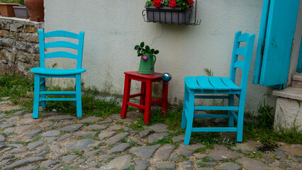 Fototapeta na wymiar Blue chairs and wooden red stool against a home wall in Gliki, an old Greek village in Gokceada. Canakkale, Turkey. There's a flower pot with red flower on the small stool table
