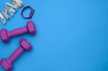 Layout of two rubberized dumbbells of 2 kg of purple color,measuring tape,fitness bracelet on a blue background,top view