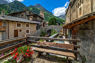 Old houses, medieval stone bridge and wooden bench in small town of Chianale, Italy.