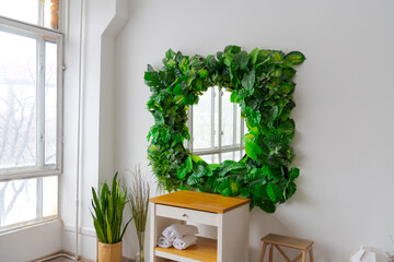Mirror with green artificial tropical leaf frame hangs above table in white bathroom. Window reflects in mirror surface. Soft focus. No people.