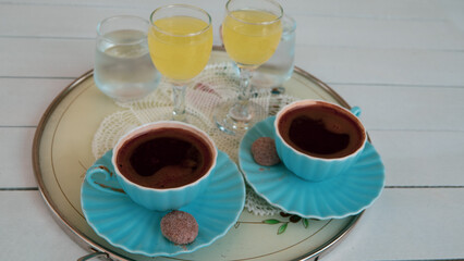 Two cups of turkish coffee served with a litle cookie and lemon liqueur or limoncello on tray on white wooden table