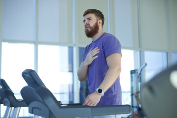 Tired exhausted young man is training workout, running on treadmill in gym holding his heart with...