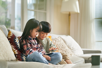 two asian children brother and sister sitting on family couch playing computer game using digital tablet