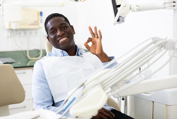 Portrait of positive african-american man sitting on dental chair, making ok gesture and smiling.